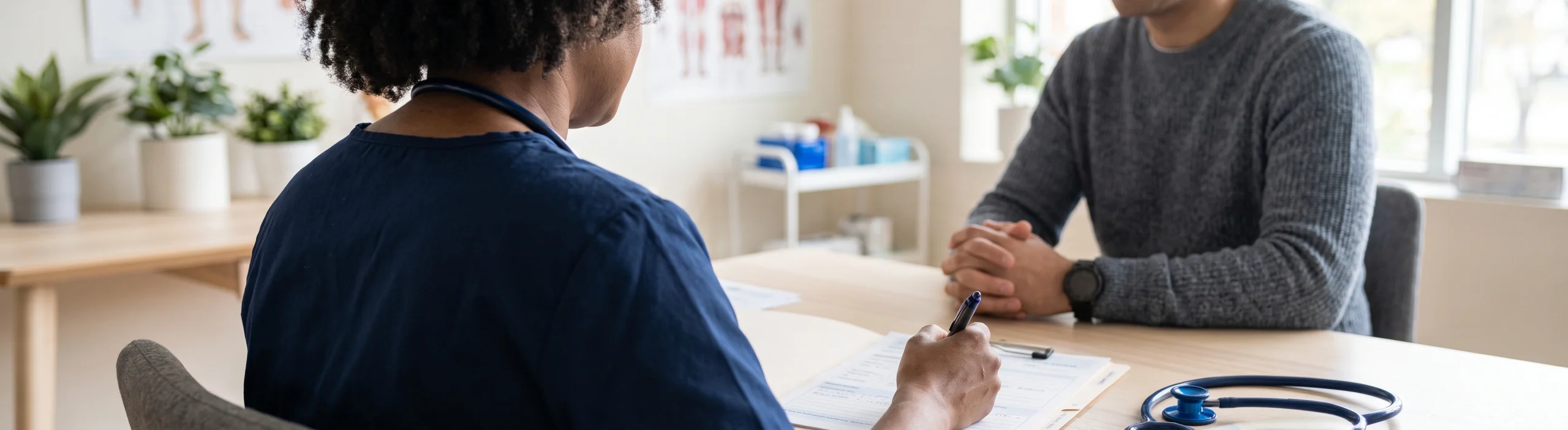 A clinician fills in an intake form while an injured patient sits across the desk, anatomical charts on the wall behind them.