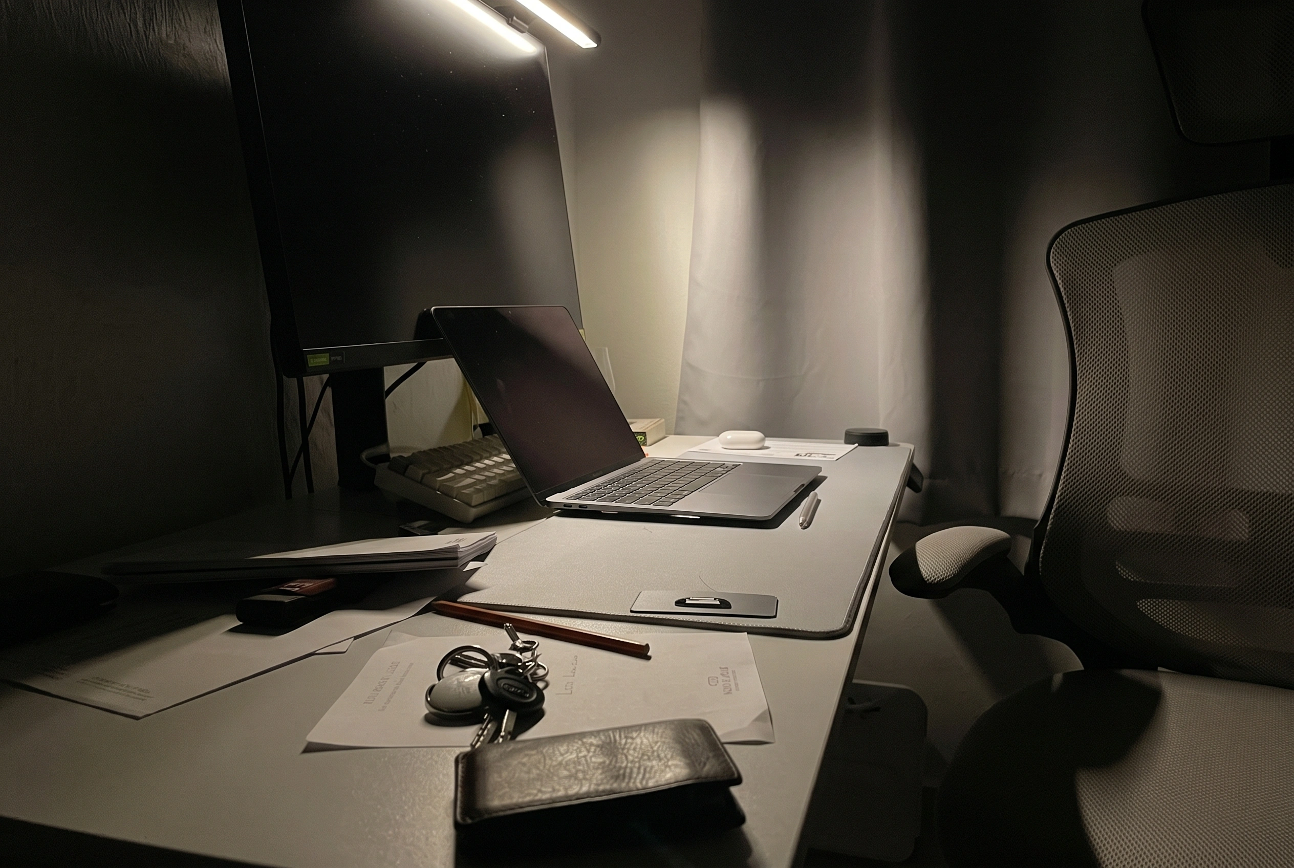 A dark room lit only by a desk lamp, with a laptop, keys and a wallet left on the desk and the screen switched off.